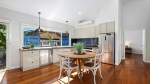 A modern kitchen with white cabinetry, a stainless steel refrigerator, and a dining area with a wooden table and white chairs. - Sawtell Beach Shack 2 (Sawtell)