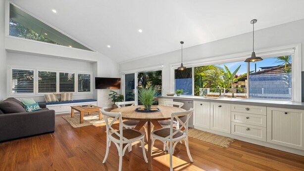A spacious kitchen with a dining area, white cabinetry, and a large window offering a view of the outdoors. - Sawtell Beach Shack 2 (Sawtell)