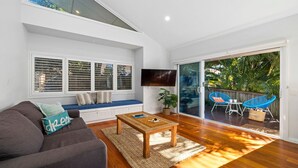 A modern living room with a sofa, a coffee table, and a TV mounted on the wall. There is a sliding glass door leading to an outdoor area with a blue chair and a plant. - Sawtell Beach Shack 2 (Sawtell)