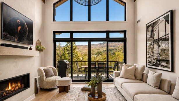 Amplia sala de estar con chimenea, asientos cómodos, un ventanal con vista a la montaña y un balcón con mobiliario exterior.