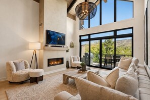 A spacious living room with a fireplace, comfortable seating, a television, and large windows offering a view of the outdoors.