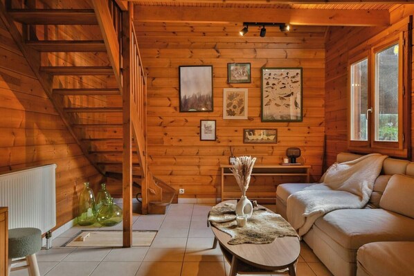 A cozy cabin interior with a wooden staircase, a sofa, a coffee table, and framed pictures on the walls. - Chalet du Schlossthal - Pond (Lemberg)