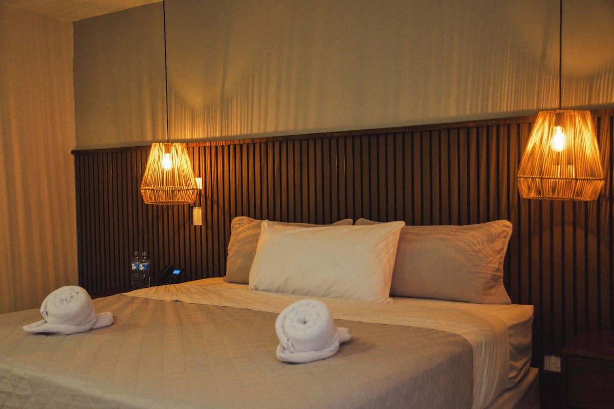 Habitación de hotel con una cama grande, dos almohadas, dos toallas enrolladas y dos lámparas fijadas a la pared.