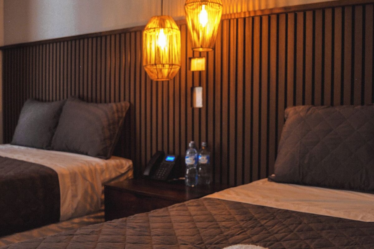 Habitación de hotel con dos camas, una mesita de noche con un teléfono y botellas de agua, y dos lámparas de pared.
