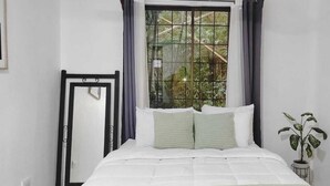 A bedroom with a bed, a window with curtains, a plant, and a mirror. - Fully Equipped Kitchen Privacy & Pura Vida (Quepos)