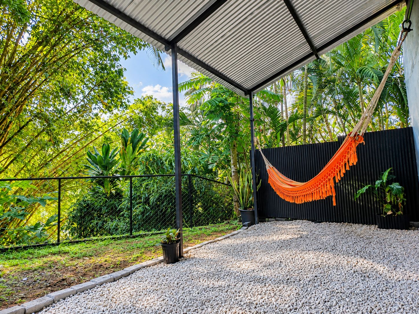An outdoor covered area with a hammock, surrounded by greenery and a black fence.
