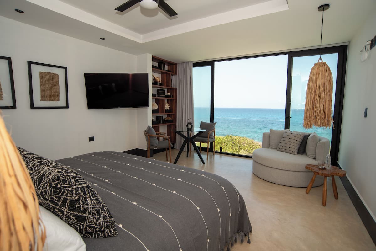 A bedroom with a large bed, a TV, a bookshelf, a seating area with a view of the ocean, and a balcony with a hanging light fixture.