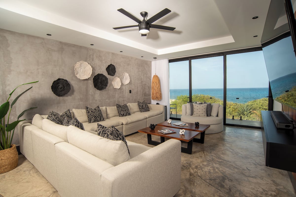 A modern living room with a sofa, coffee table, and a television. There is a large window with a view of the ocean.