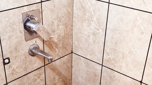 Une salle de bain avec des carreaux de couleur beige, des toilettes, une douche et un récipient en plastique posé sur le sol.