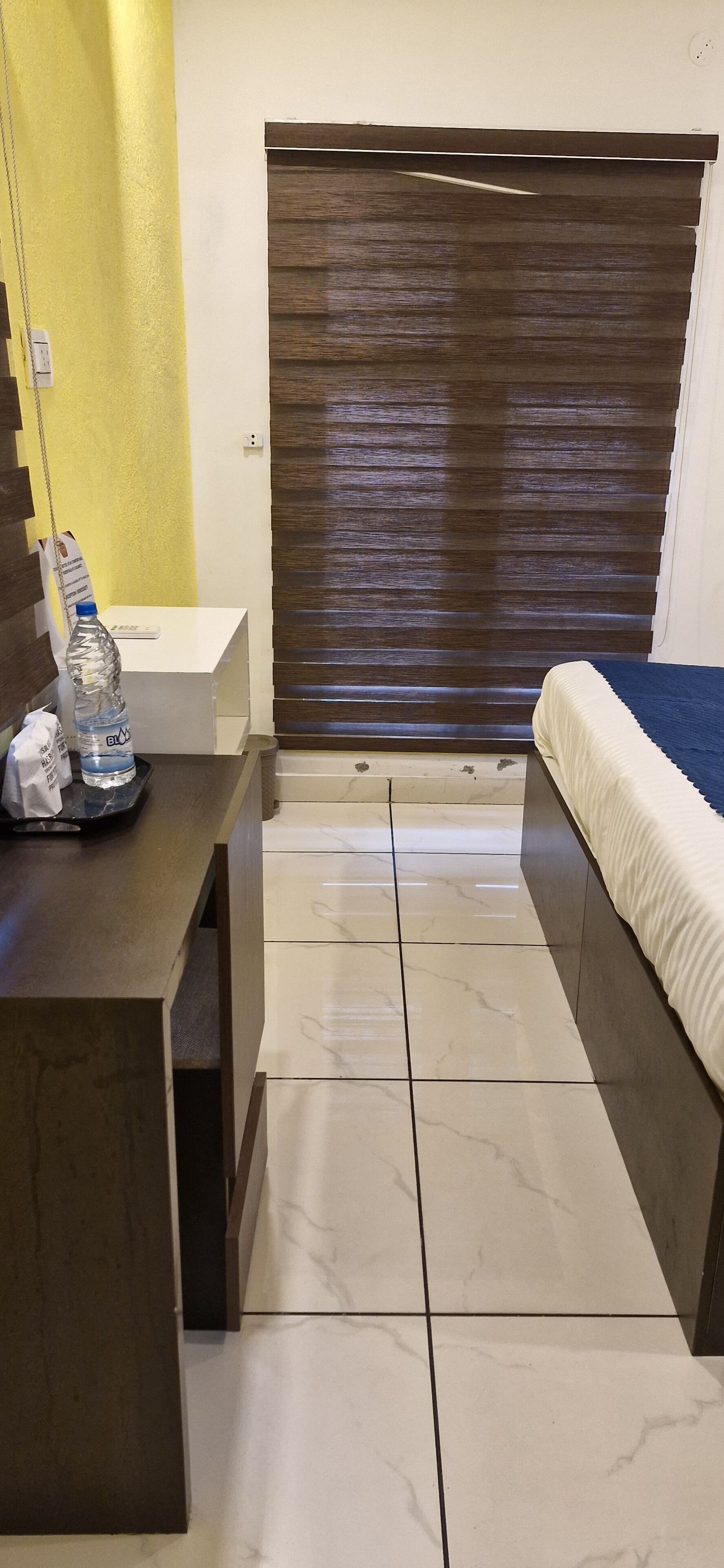 A hotel room with a bed, a desk, a chair, a water bottle, a tissue box, and a window with blinds.