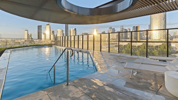 An outdoor swimming pool with a city skyline view, surrounded by a modern architectural structure with a circular design element. - Zi apartments - Balcony, Bayview, Sauna & Pool (South Melbourne)