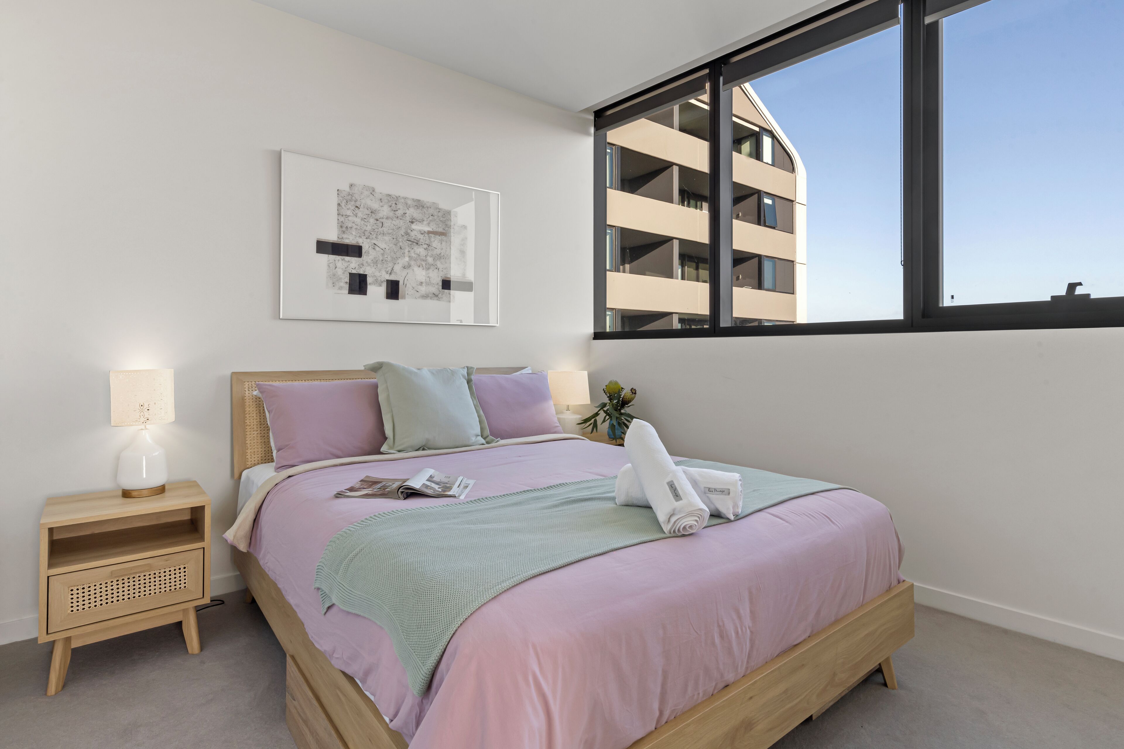 A modern bedroom with a large window, a wooden bed, a bedside table with a lamp, and a framed artwork on the wall.