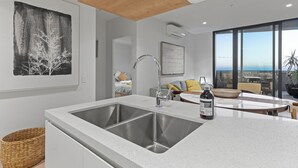 A modern kitchen with a large island, stainless steel sink, and a dining area with a view of the ocean. - Zi apartments - Balcony, Bayview, Sauna & Pool (South Melbourne)