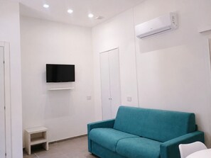 Une pièce avec un canapé de couleur turquoise, un téléviseur posé sur une étagère blanche, une porte blanche et un appareil de climatisation fixé au mur.