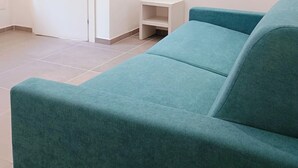 A teal sofa, a white door, and a mounted TV in a room with white walls and tiled flooring.