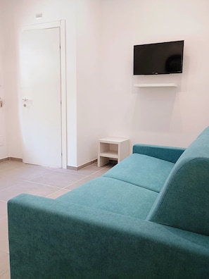 A teal sofa, a white door, and a mounted TV in a room with white walls and tiled flooring.