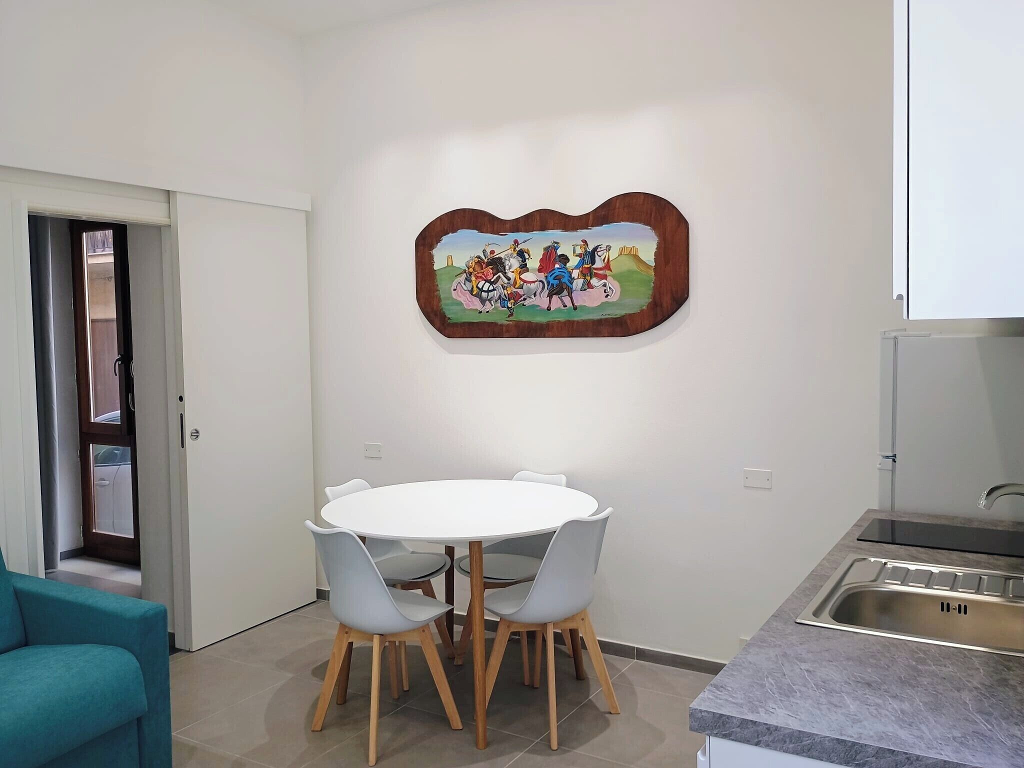 Ein Essbereich mit rundem Tisch und Stühlen, eine Küche mit Spüle aus Edelstahl und ein an der Wand befestigtes Kunstwerk.