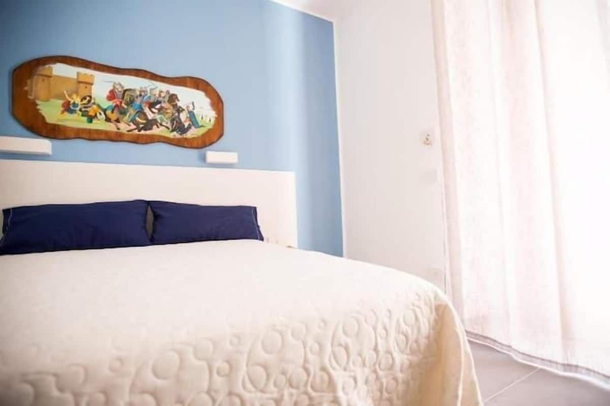 Een slaapkamer met een bed, een schilderij aan de muur en een raam met gordijnen.