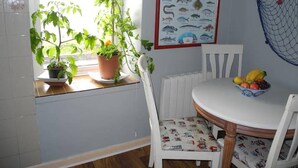 Een keuken met een raam, kamerplanten, een visillustratie op de muur, een witte stoel en een tafel met fruit.