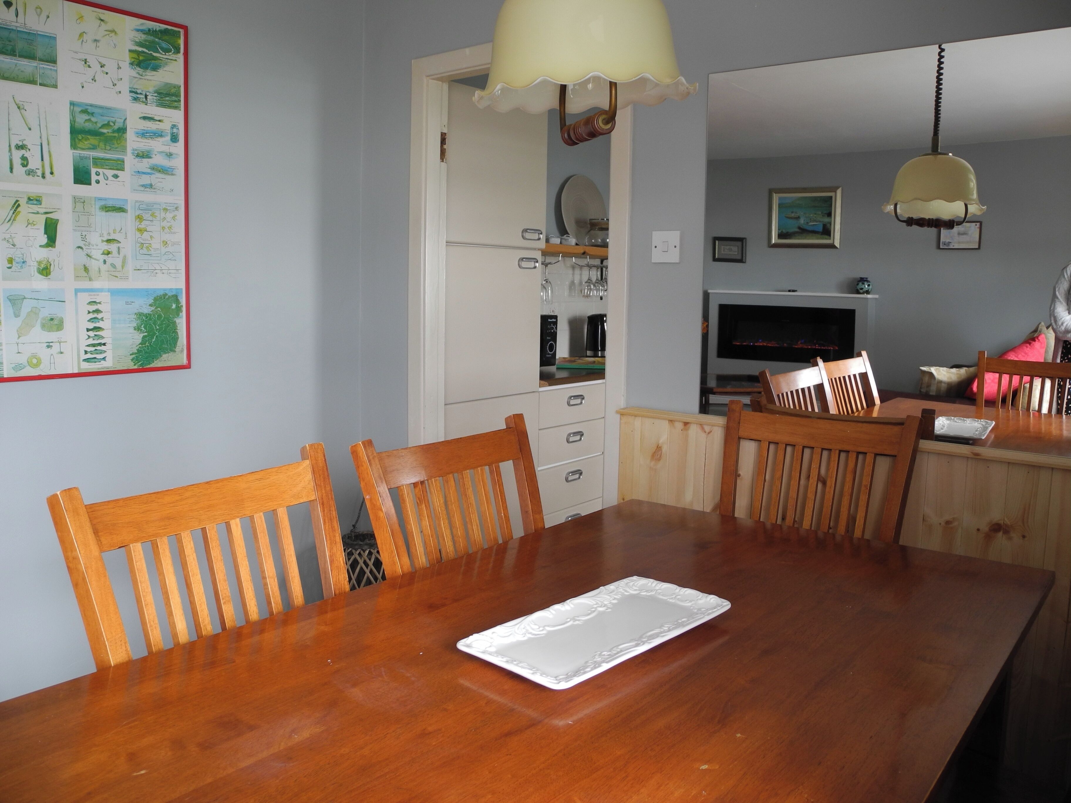 A dining area with a wooden table and chairs, a white placemat, and a kitchen in the background with a TV and various items on the counter.