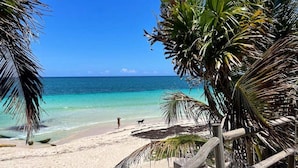 Een strand met helder turquoise water, een zandstrand en een houten terras met zitplaatsen.