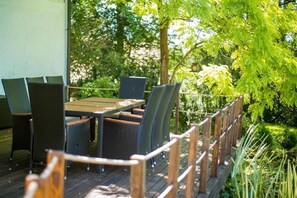 Een terras buiten, met een houten tafel en rieten stoelen, omgeven door een houten balustrade en weelderige beplanting.