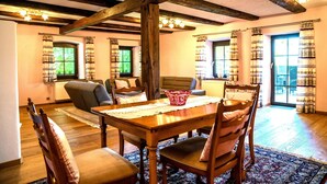 A dining area with a wooden table, chairs, and a chandelier. There are windows with curtains and a view of the outdoors. - Vacation home in the district of Passau with plenty of space and comfort (Windorf)