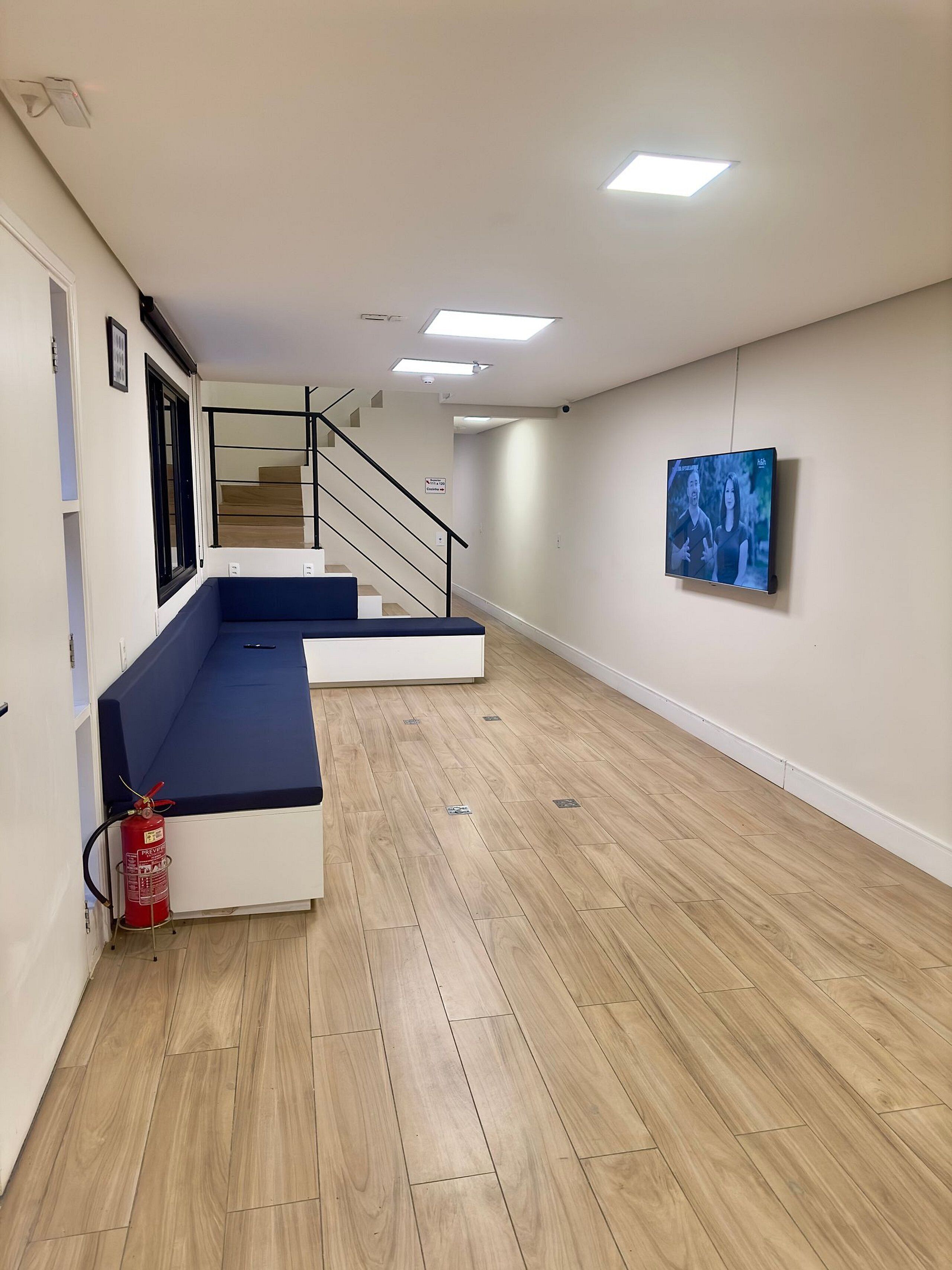 Una habitación amplia con piso de madera, un sofá azul, una escalera y un televisor montado.