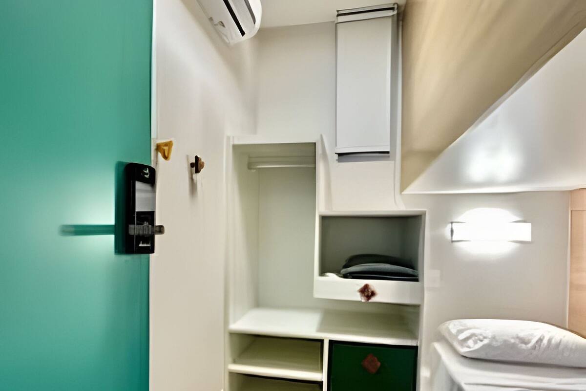 A compact room with a bunk bed, a built-in wardrobe, a small seating area, and a wall-mounted air conditioning unit.
