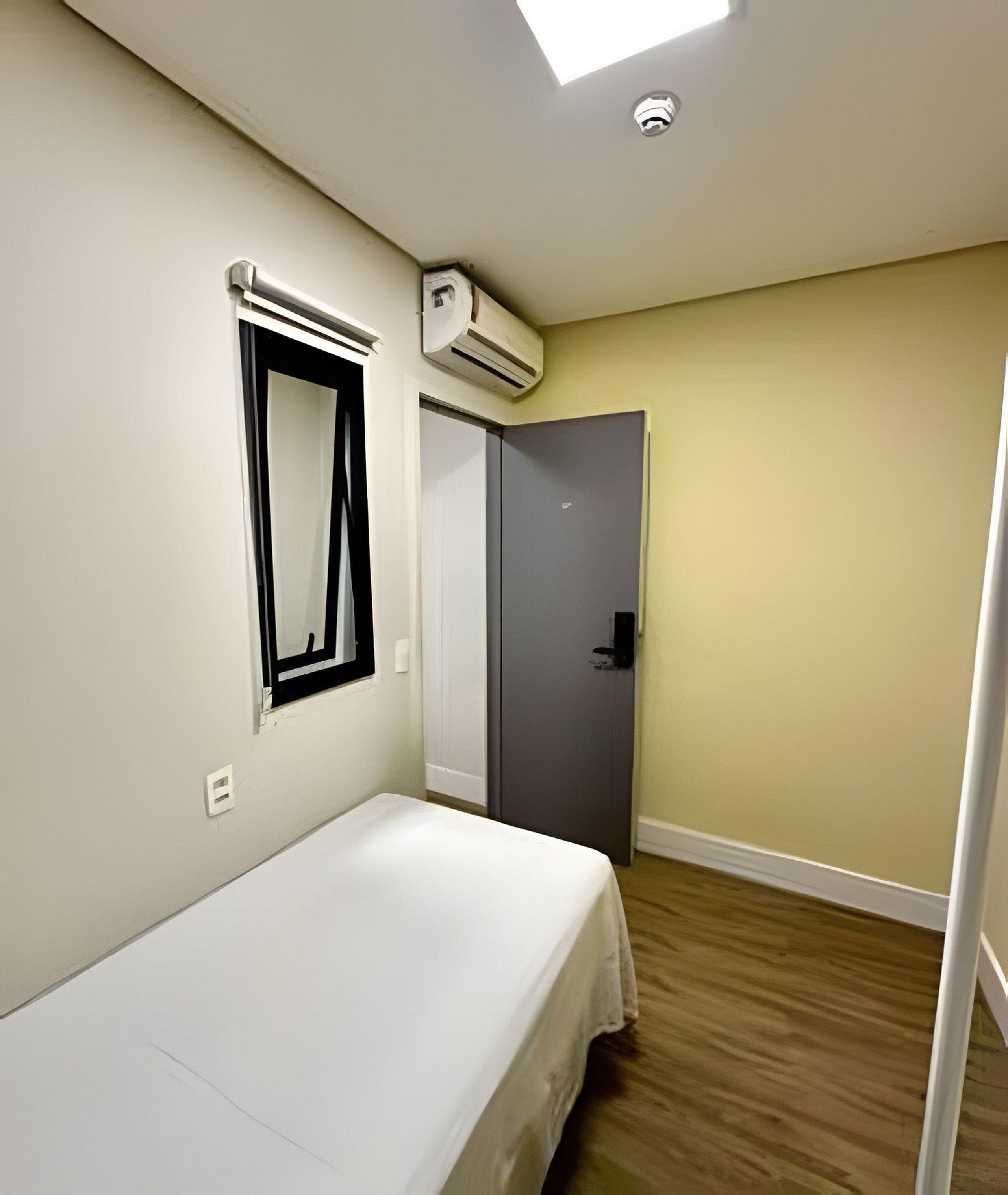 A room with a bed, an air conditioning unit, a mirror, a door, and a light fixture.