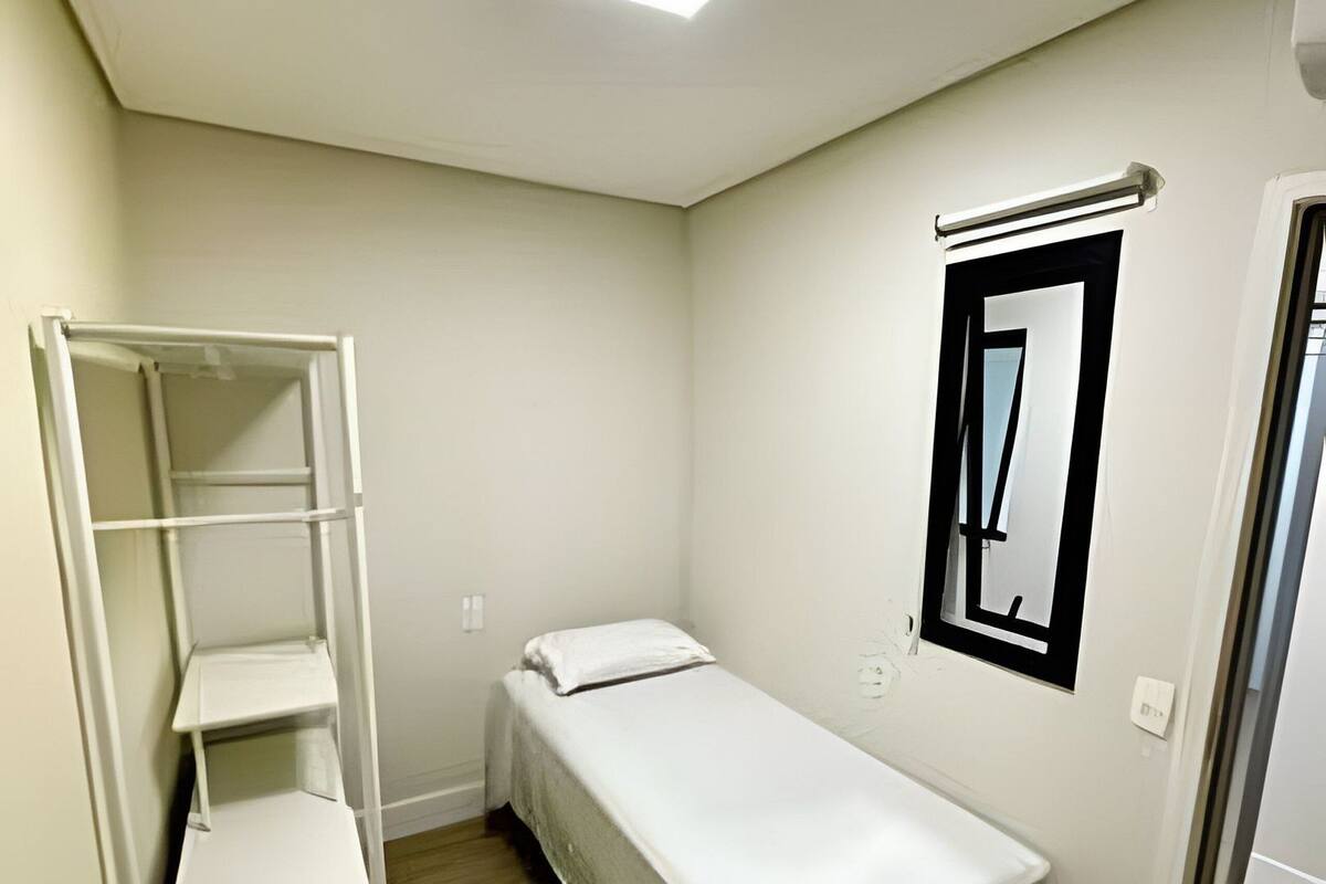 A small, single-bed room with a white bed, a white shelf unit, a ceiling light, and a door.