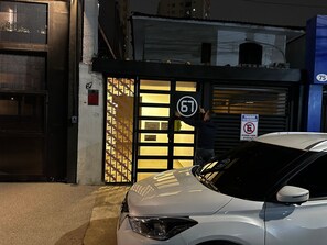 A building entrance with a numbered gate, a car parked in front, and a no parking sign. - Quartos Vila Olimpia  (São Paulo)