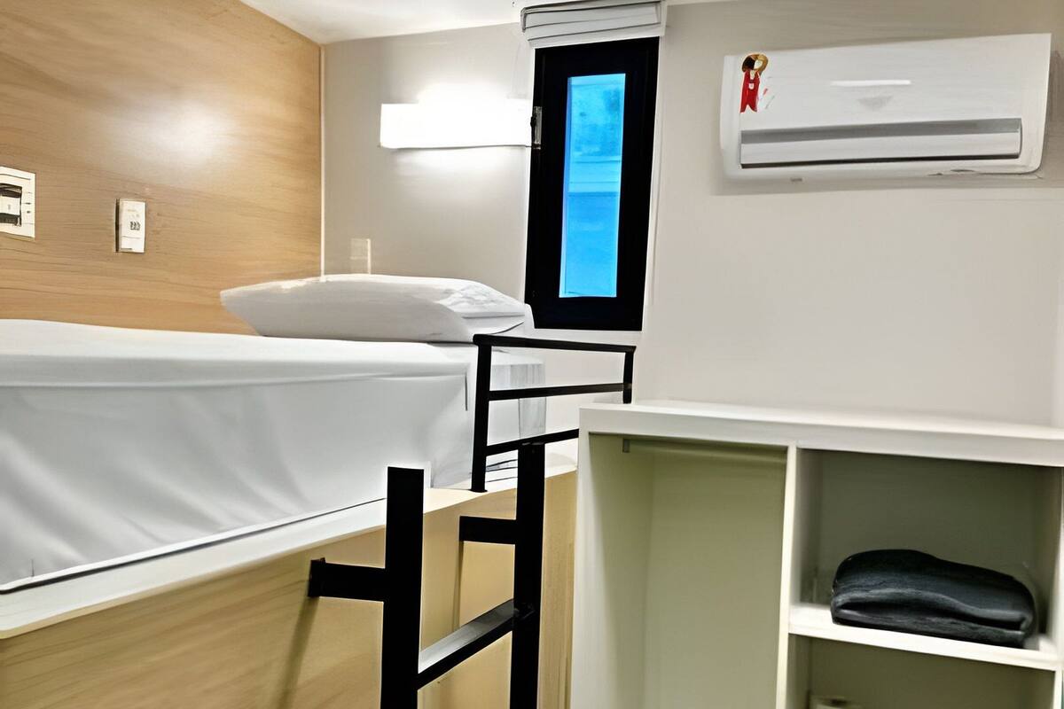 A bunk bed with a ladder, a desk with a chair, a shelf with folded clothes, and an air conditioning unit.
