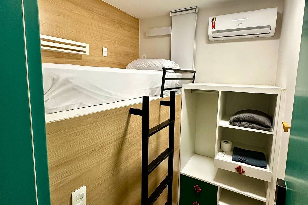 A compact room with a bunk bed, a desk with a chair, a shelf with storage, and an air conditioning unit.