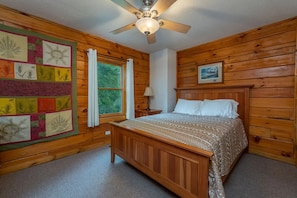 A wooden-paneled room with a bed, a quilt, a ceiling fan, and a window.