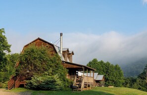 Une grange rustique en bois, dotée d’une cheminée, entourée d’une végétation luxuriante et sous un ciel dégagé.