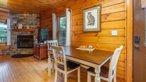 Interior de una cabaña de madera con chimenea de piedra, una mesa de comedor con sillas, un televisor y una imagen de un búho enmarcada en la pared.