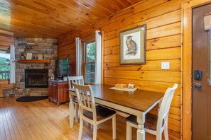 Interior de una cabaña de madera con chimenea de piedra, una mesa de comedor con sillas, un televisor y una imagen de un búho enmarcada en la pared.