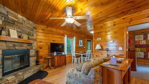 A cozy cabin interior with a stone fireplace, wooden walls, a ceiling fan, a television, a floral sofa, and a dining area. - Mt. Mitchell Log Cabin | Engadine Inn & Cabins (Candler)