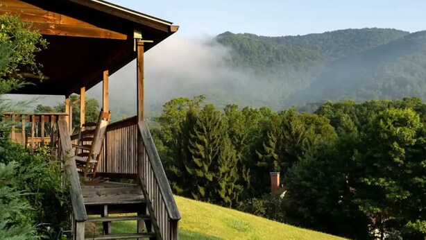 Una cabaña de madera con un porche que da a un valle boscoso y colinas cubiertas de niebla.