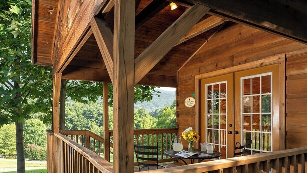 Een houten hut met een terras, een tafel met stoelen en uitzicht op een bebost berglandschap.