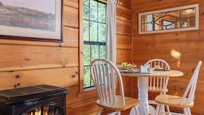 L’intérieur d’un chalet en bois chaleureux et accueillant, avec une cheminée, des chaises en bois peintes en blanc, une table à manger où sont disposés des aliments pour le petit-déjeuner, et un grand tableau représentant un paysage, encadré.