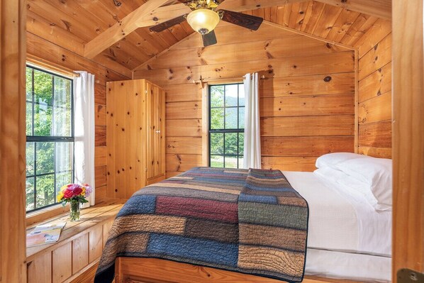 A wooden cabin bedroom with a bed, a quilt, a window, and a ceiling fan.