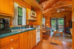 A cozy cabin interior with wooden walls, a kitchen with a stove and microwave, a dining area with a table and chairs, and a large window overlooking greenery.