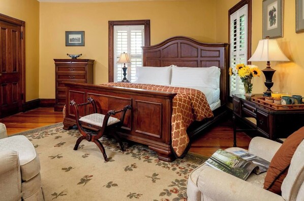A bedroom with a wooden bed, a wooden nightstand with a lamp, a chair, a dresser, and a floral carpet. - Vineyard View Suite | Engadine Inn & Cabins (Candler)