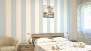 Une chambre à coucher avec un lit, une table de chevet et une chaise. Un mur est décoré de rayures verticales, et un tableau encadré est accroché au-dessus du lit.