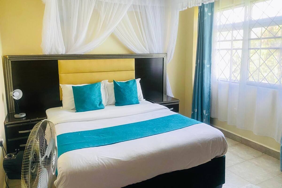 Une chambre d’hôtel avec un lit, une table de chevet, un ventilateur et une fenêtre avec des rideaux.