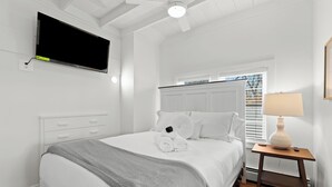 Un dormitorio con cama, televisor montado en la pared, mesita de noche con lámpara y ventana con persianas.