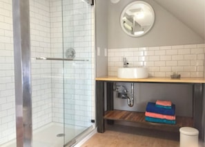 Un baño con cabina de ducha de vidrio, un lavamanos blanco y un estante de madera con toallas.