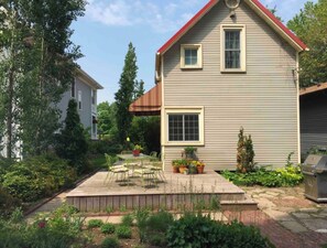 Una casa de dos pisos con techo rojo, un porche con mesa y sillas, y un jardín con diversas plantas y flores.